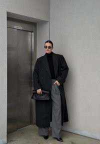 Long manteau en laine noire à double boutonnage, pantalon gris à jambes larges, tenant une pochette en cuir noire et portant des lunettes de soleil foncées.