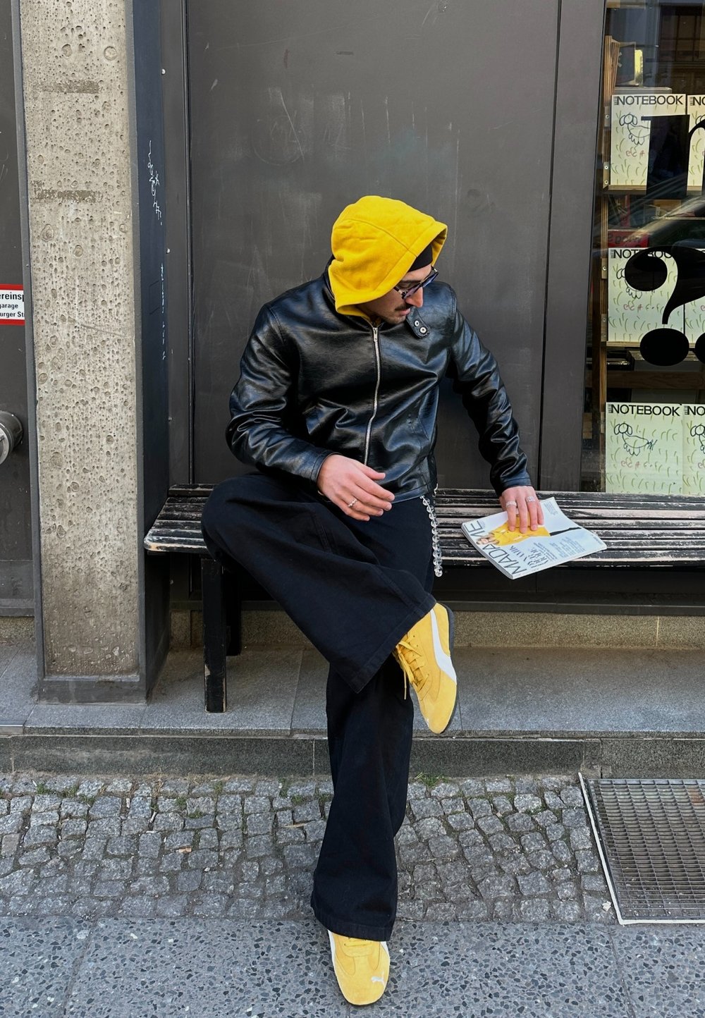 Μαύρο δερμάτινο μπουφάν με κίτρινη κουκούλα, μαύρο παντελόνι σε φαρδιά γραμμή και κίτρινες αθλητικές γόβες. Καθισμένος σε ξύλινο παγκάκι, κρατώντας ένα περιοδικό. Σκηνικό από σκυρόδεμα.