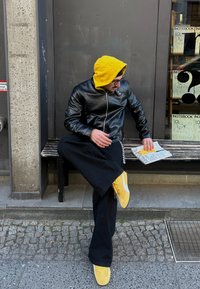 Μαύρο δερμάτινο μπουφάν με κίτρινη κουκούλα, μαύρο παντελόνι σε φαρδιά γραμμή και κίτρινες αθλητικές γόβες. Καθισμένος σε ξύλινο παγκάκι, κρατώντας ένα περιοδικό. Σκηνικό από σκυρόδεμα.