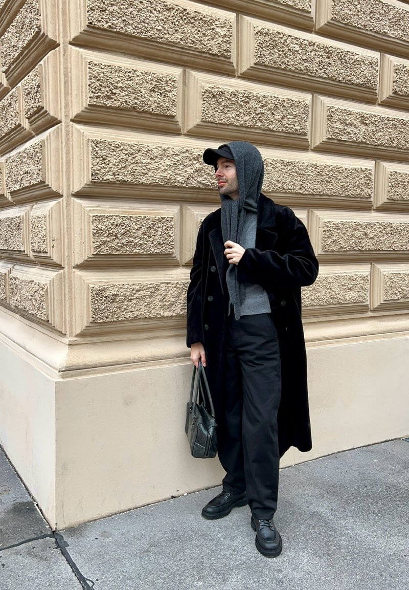 Cappotto lungo in velluto nero, felpa con cappuccio grigia, sciarpa grigia, pantaloni neri, scarpe nere, tiene una borsa scura, in piedi contro un muro beige testurizzato.