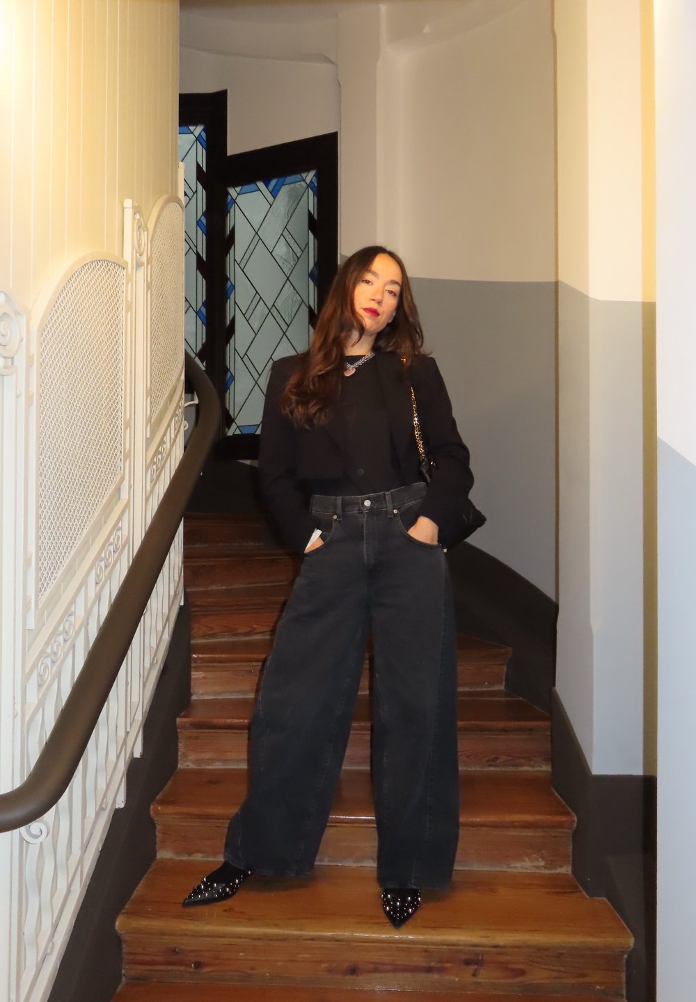 Zwart cropped jack over een zwart topje, gecombineerd met wijde zwarte jeans. Glimmende zwarte pumps met puntige neus, staand op houten trappen met wanddetails.