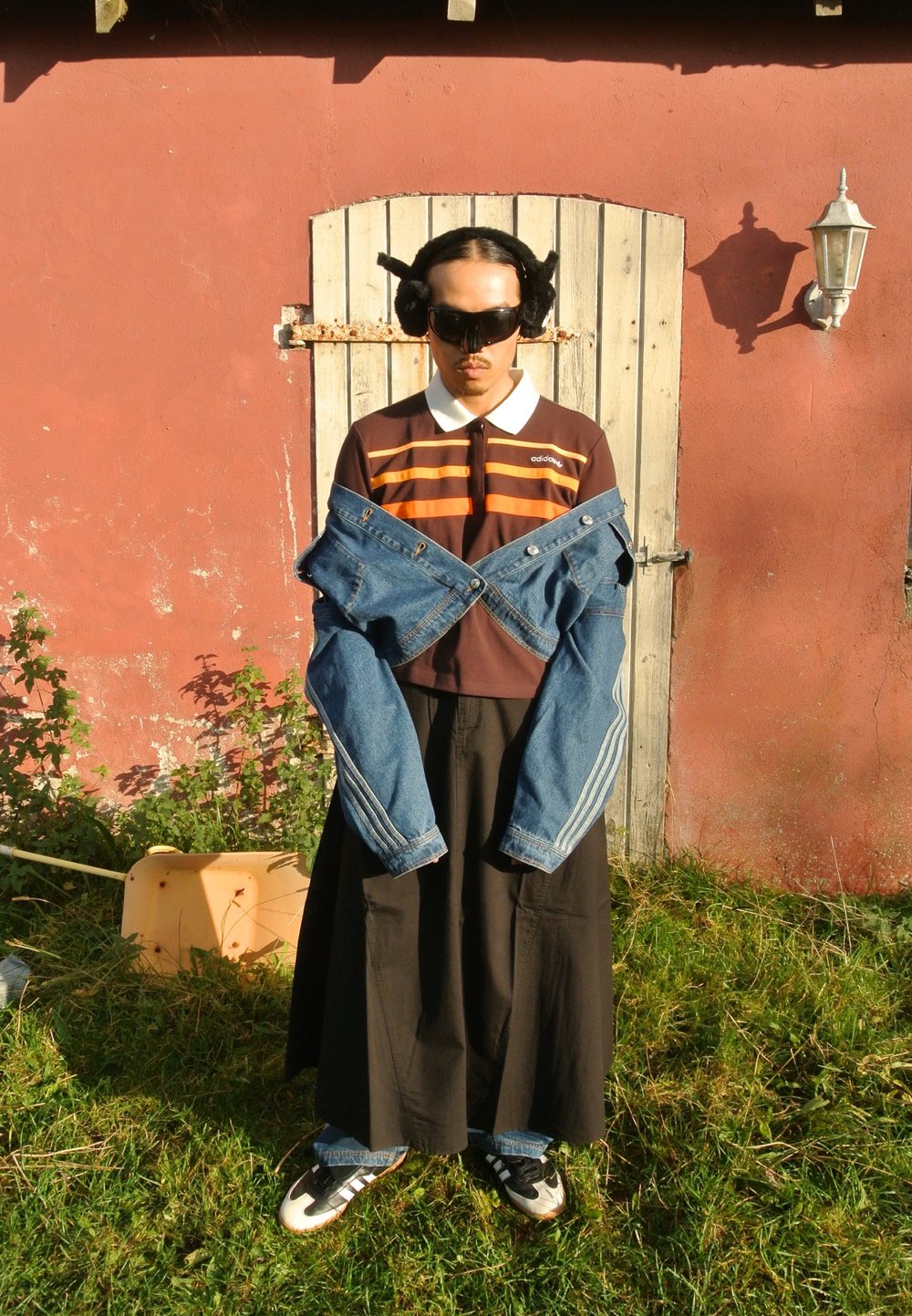 Ένα άτομο φοράει μια σκούρα φούστα και ένα καφέ πόλο με πορτοκαλί ρίγες, κρατώντας μια τζιν ζακέτα. Το φόντο περιλαμβάνει μια ξύλινη πόρτα και πράσινα φυτά.