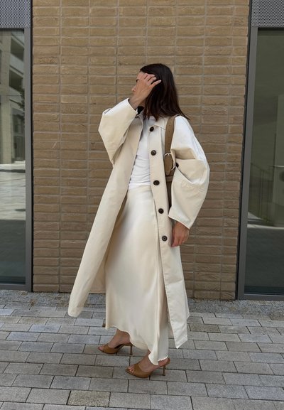 Abrigo trench beige con botones grandes, llevado sobre una blusa blanca y una falda de satén. El conjunto incluye un bolso de hombro con bandolera, combinado con tacones marrones.