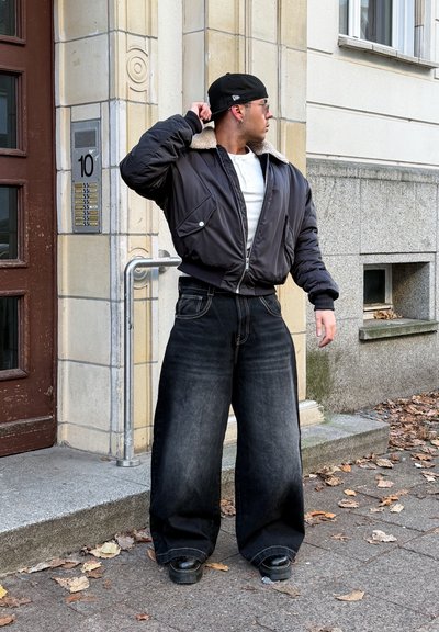Chaqueta bomber negra con cuello de borrego, camisa blanca y jeans oscuros de gran tamaño. Zapatillas negras completan el conjunto. Fondo urbano con vegetación.