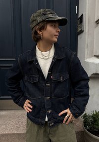 Osoba nosící maskáčovou čepici, tmavě modrou džínovou bundu, bílý svetr a olivové kalhoty stojí s rukama v bok a dívá se doprava před budovou.