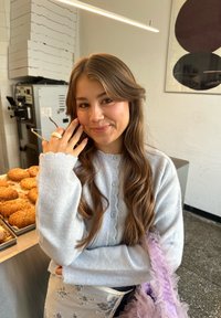 Cardigan grigio chiaro con colletto arricciato, abbinato a una borsa viola trasparente e con volant. Sullo sfondo, prodotti da forno e attrezzature da cucina industriale.