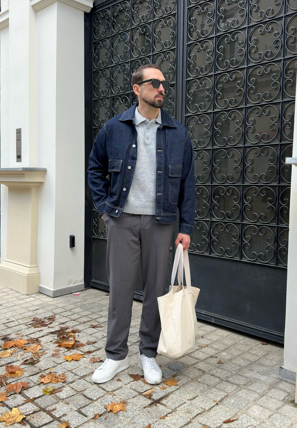 Mężczyzna w okularach przeciwsłonecznych, w dżinsowej kurtce, szarym swetrze i spodniach, trzymający beżową torbę na zakupy, stoi na brukowanej chodniku pokrytym jesiennymi liśćmi.