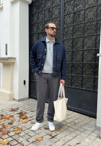 Mężczyzna w okularach przeciwsłonecznych, w dżinsowej kurtce, szarym swetrze i spodniach, trzymający beżową torbę na zakupy, stoi na brukowanej chodniku pokrytym jesiennymi liśćmi.