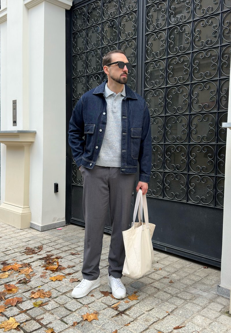 Mann trägt Sonnenbrille, Jeansjacke, grauen Pullover und Hose, hält eine beigen Baumwolltasche und steht auf einem Kopfsteingehweg mit Herbstblättern.