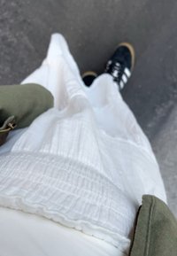 Bílá strukturovaná sukně s volánovým lemem, kombinovaná s černými sportovními botami s bílými pruhy. Obsahuje detail olivově zelené bundy.