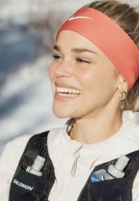 Femme souriante portant un bandeau Nike orange, une veste blanche et un gilet d'hydratation Salomon noir avec des bouteilles d'eau dans les poches.