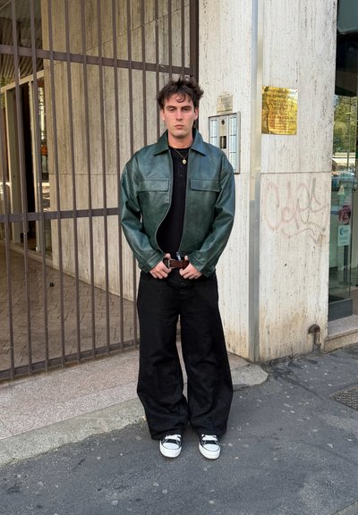 Joven con chaqueta de cuero verde, camisa negra, pantalones anchos negros y zapatillas, apoyado contra un edificio con puerta metálica y grafiti.