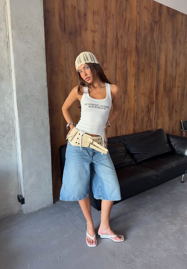 Jeune femme portant un débardeur blanc avec texte, un short en jean, un bonnet tricoté et des sandales, posant à l'intérieur près d'un canapé noir et d'un mur en bois.