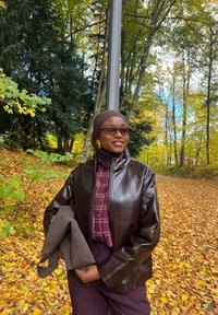Veste en cuir synthétique marron, chemise à carreaux bordeaux, pantalon marron, tenant un accessoire en tissu marron. Entouré de feuillage d'automne et d'arbres.