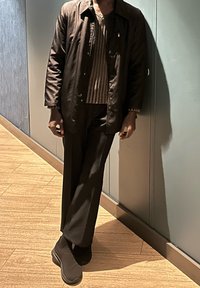 Άτομο που φοράει σκούρο μπουφάν, ριγέ πουκάμισο, μαύρο παντελόνι και μαύρα αθλητικά παπούτσια, στέκεται μέσα σε εσωτερικό χώρο πάνω σε ξύλινο πάτωμα, μπροστά σε γκρι τοίχο.