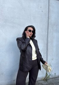 Brown leather blazer over a cream top, paired with dark pants. Holding a light cream handbag. Black sunglasses complete the look.