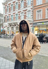 Homme portant une veste à capuche beige et une casquette noire debout dans une rue de la ville avec un bâtiment historique et un magasin Holland & Barrett en arrière-plan.