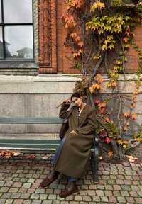Person i en stor olivengrønn frakk sitter på en grønn benk med en brun veske, omgitt av røde og gule blader foran en murvegg.