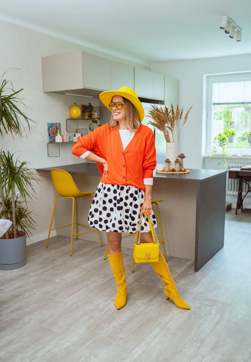 Cardigan arancione, maglietta bianca, gonna a pois, stivali gialli fino al ginocchio e borsa coordinata. Colori vivaci e varie trame in un ambiente cucina moderno.