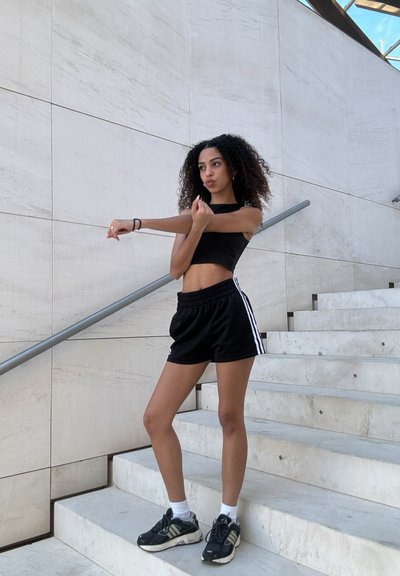 Top corto negro y pantalones cortos negros con rayas blancas, combinados con zapatillas deportivas negras, de pie sobre escalones de piedra blanca.