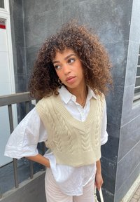 Maglione senza maniche a treccia color beige con scollatura a V, indossato sopra una camicia bianca con colletto, abbinato a pantaloni a righe di colore chiaro.
