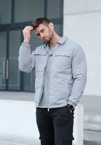 Veste matelassée gris clair avec fermeture éclair et deux poches avant, portée sur un t-shirt gris, associée à un pantalon cargo noir.