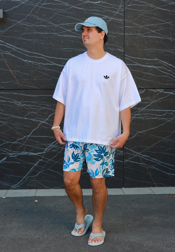 Mężczyzna w białej, oversize koszulce Adidas, niebieskich szortach w kwiaty, jasnoniebieskiej czapce i beżowych klapkach stojący przy czarnej marmurowej ścianie.