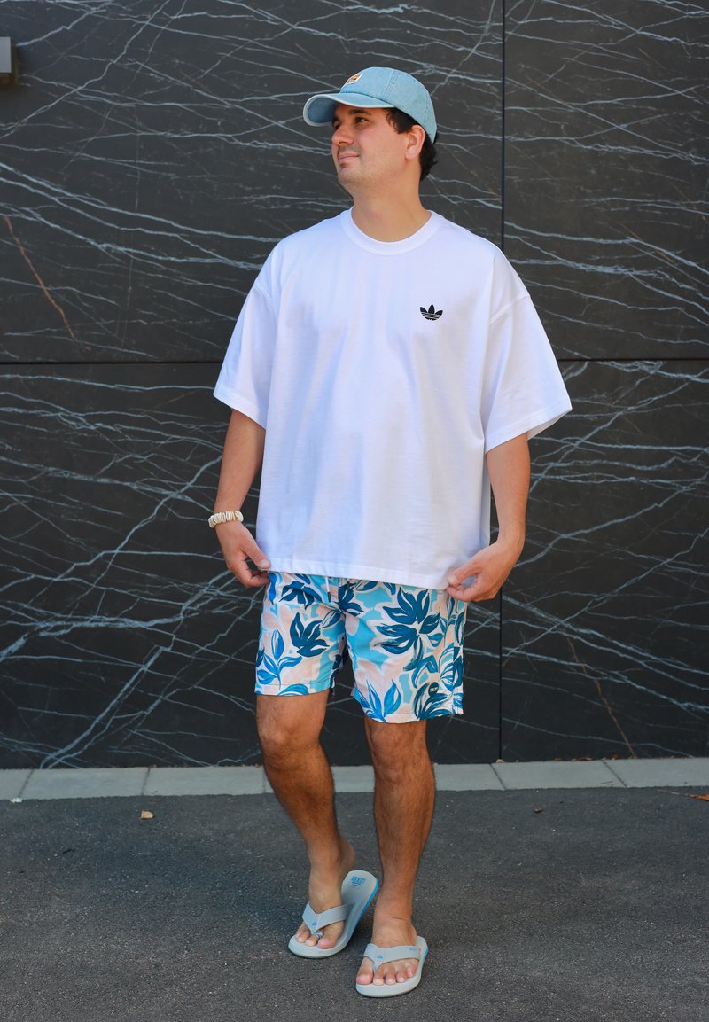 Homme portant un T-shirt Adidas blanc oversize, un short bleu à motifs floraux, une casquette bleu clair et des tongs beige, debout contre un mur en marbre noir.