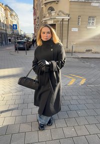 Musta villakangastakki, jossa korkea kaulus, suuret napit ja sivutaskut. Mustat nahkahansikkaat ja pieni musta krokotiilikuviollinen käsilaukku. Leveälahkeiset farkut.