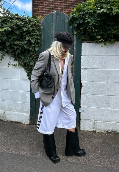 Blazer gris de gran tamaño sobre una camisa blanca, pantalones cortos blancos con rayas negras y botas de goma negras hasta la rodilla, acompañados de un bolso negro.