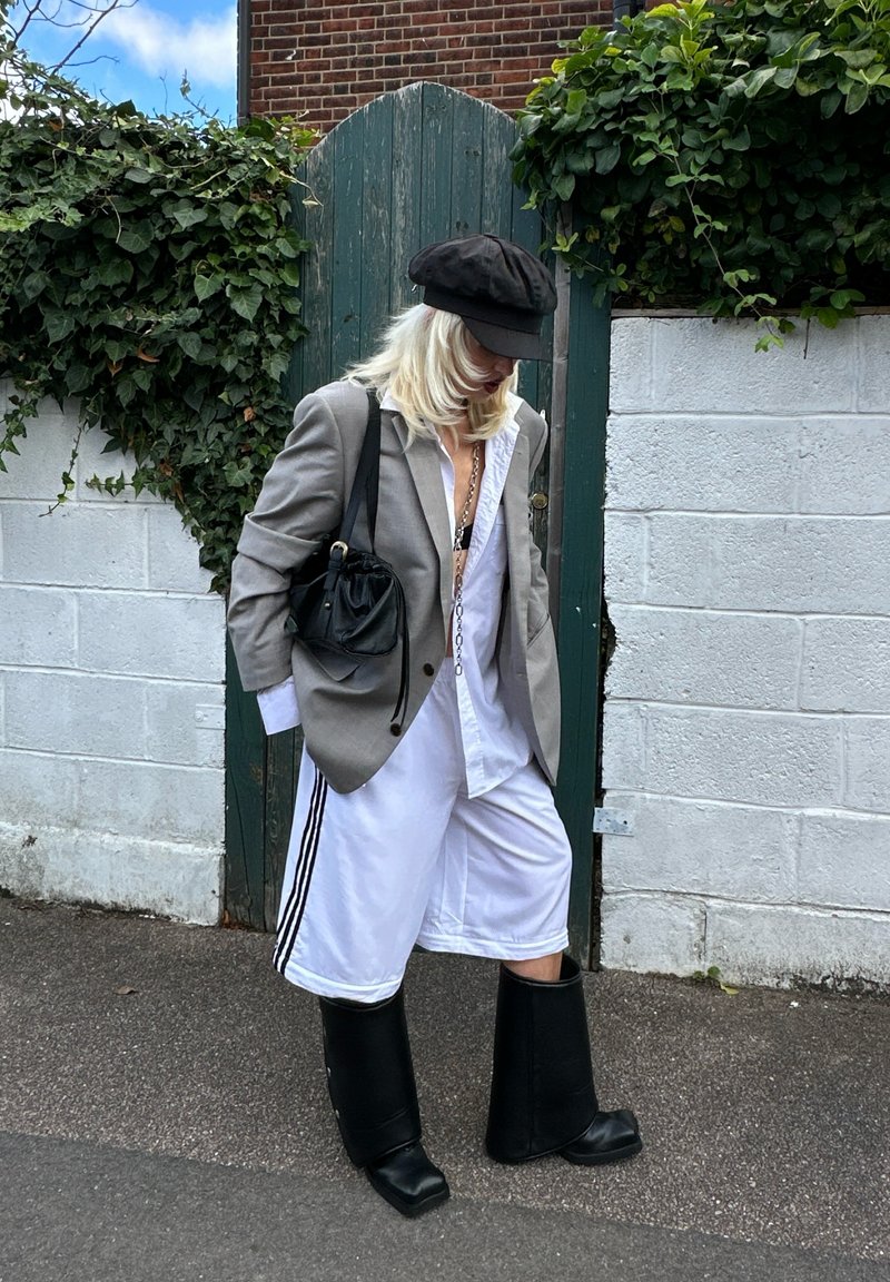 Oversized grauer Blazer über einem weißen Hemd, weiße Shorts mit schwarzen Streifen und knielangen schwarzen Gummistiefeln, kombiniert mit einer schwarzen Handtasche.