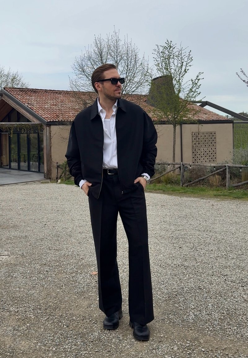 Homme vêtu d'une veste noire, chemise blanche, pantalon noir et lunettes de soleil, debout sur du gravier devant un bâtiment rustique avec des arbres en arrière-plan.