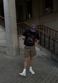 Młody mężczyzna stoi na wybrukowanej ulicy obok schodów, nosząc czapkę, czarną koszulkę z grafiką, beżowe szorty, białe skarpetki i białe trampki.