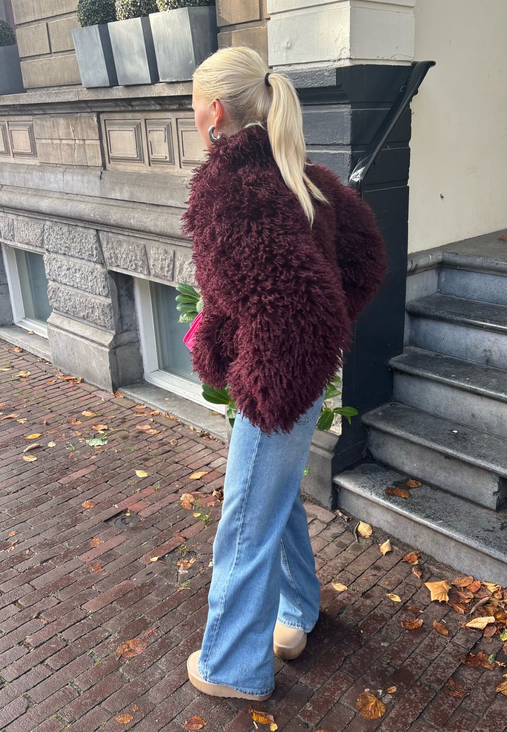 Pessoa com um cabelo loiro preso em um rabo de cavalo, vestindo uma jaqueta shaggy de um tom escuro de borgonha, jeans de perna larga em azul claro e sapatos bege, caminhando sobre uma calçada de tijolos.