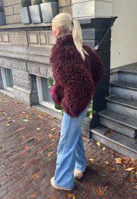 Pessoa com um cabelo loiro preso em um rabo de cavalo, vestindo uma jaqueta shaggy de um tom escuro de borgonha, jeans de perna larga em azul claro e sapatos bege, caminhando sobre uma calçada de tijolos.