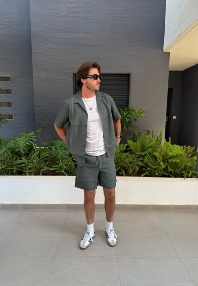 Camisa de manga corta a rayas verde con pantalones cortos a juego, llevada sobre una camiseta blanca. Combinada con gafas de sol y zapatillas blancas.