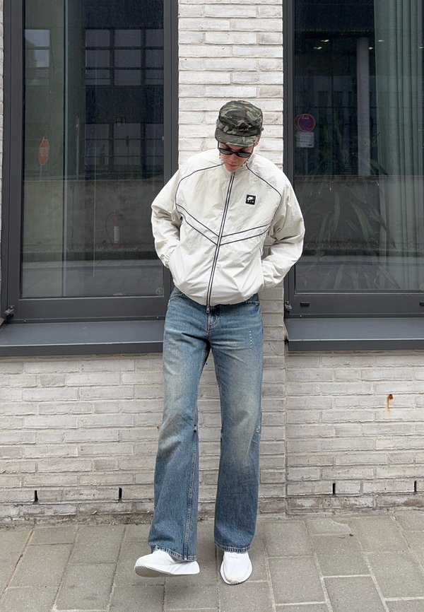 Person in weisser Reissverschlussjacke, blauer Jeans, weissen Schuhen, Camouflage-Hut und Sonnenbrille, stehend mit Händen in den Taschen vor einer weissen Backsteinmauer.
