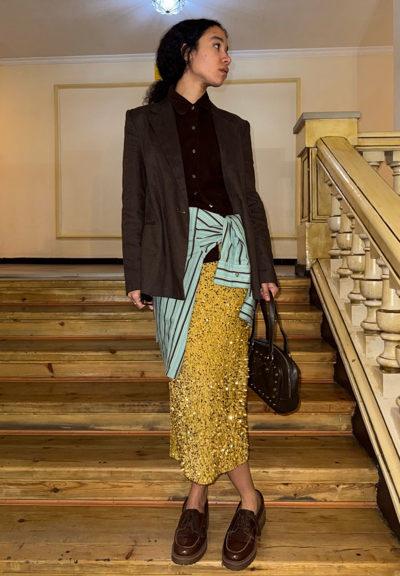 Jeune personne debout sur des escaliers en bois portant un blazer sombre, une chemise rayée nouée à la taille, une jupe en sequins dorés, des chaussures marron, et tenant un sac à main.