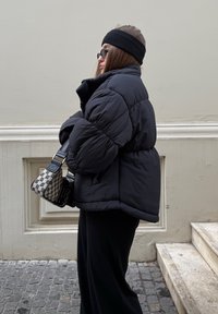 Giacca a vento imbottita nera con collo alto, indossata sopra pantaloni neri. Presenta una borsa con motivo decorativo e una fascia nera per capelli. Lo sfondo è una parete di colore chiaro.