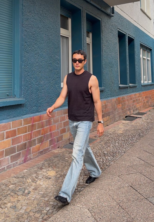 Man met een zwarte mouwloze shirt, lichtblauwe jeans, zwarte schoenen en een zonnebril die op een kasseienstoep naast een blauw bakstenen gebouw loopt.