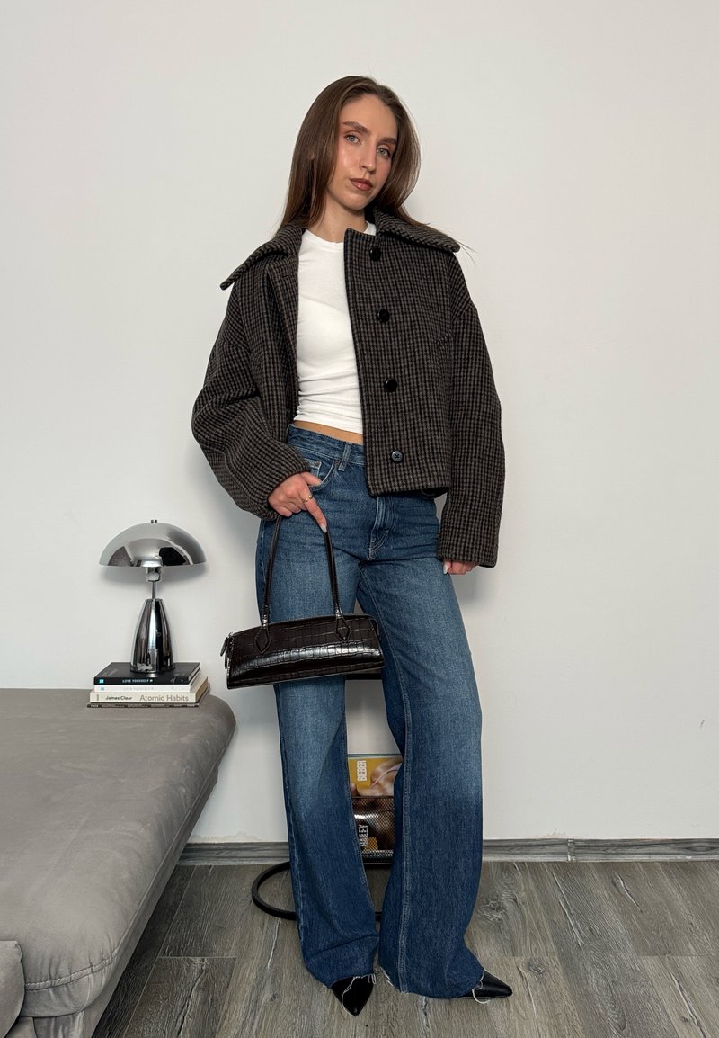 Giovane donna con giacca corta a quadri, top bianco, jeans a gamba larga e scarpe nere a punta, che tiene una borsa nera in un ambiente interno.