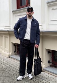 Veste marine avec deux poches avant portée sur une chemise blanche, pantalon noir et baskets argentées ; tenant un sac fourre-tout noir.