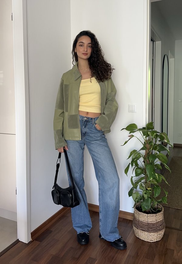 Femme aux cheveux bouclés portant une veste verte, un crop top jaune, un jean bleu ample, des chaussures noires, tenant un sac à main noir, debout près d'une plante d'intérieur dans un panier tressé.