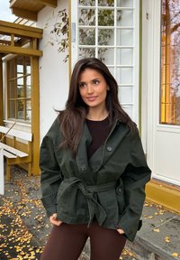 Giovane donna con lunghi capelli castani che indossa una giacca verde scuro con cintura e pantaloni marroni, in piedi davanti a un edificio giallo e bianco.