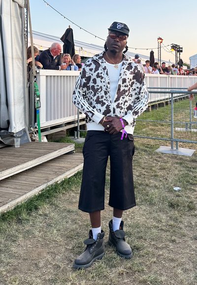 Chaqueta de cremallera con estampado de vaca en blanco y negro, camiseta blanca, pantalones cortos negros y botas negras con detalles grises. Ambiente al aire libre con personas al fondo.