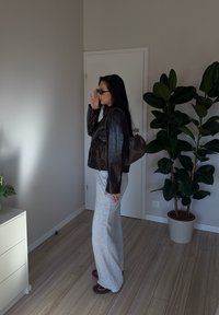 Žena v čiernej koženej bunde, šedých širokých nohaviciach a slnečných okuliaroch, ktorá si upravuje okuliare vo vnútri, stojí vedľa veľkej kvetináča pri bielych dverách.