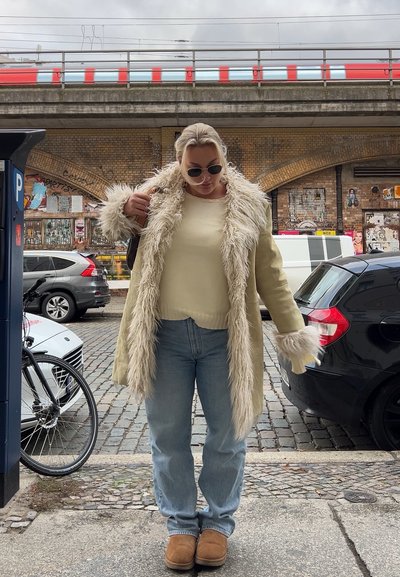 Abrigo de piel sintética beige con mangas largas, suéter de punto color crema, jeans de mezclilla azul claro y zapatos tipo slip-on color marrón, de pie en una calle de adoquines.