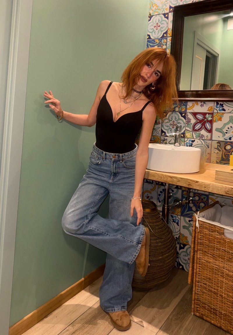 Jeune personne aux cheveux roux portant un débardeur noir et un jean large, appuyée contre un mur vert près d'un lavabo de salle de bain décoré et d'un panier à linge en osier.