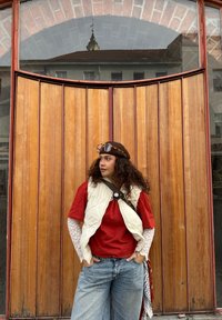 Persona sarkānā kreklā, krēmkrāsas jaku un gaiši ziliem platajiem džinsiem, ar cirtainiem matiem un brillēm, stāv pie koka durvīm.