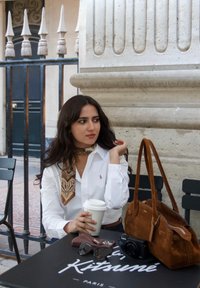 Chemise blanche boutonnée avec une écharpe à motif cachemire, tenant une tasse de café. Un sac à main en daim marron et des lunettes de soleil posés sur une table noire avec un logo.